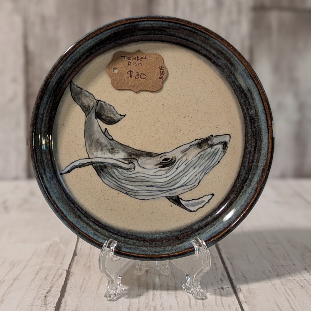 Small handmade ceramic plate. There is an underglaze painting of a humpback whale in greys and grey-browns on white clay. The rim is glazed with blue Rutile, a deep blue glaze that breaks a brown color at the rim. The plate is propped up on a stand against a white and grey faux wood background. 