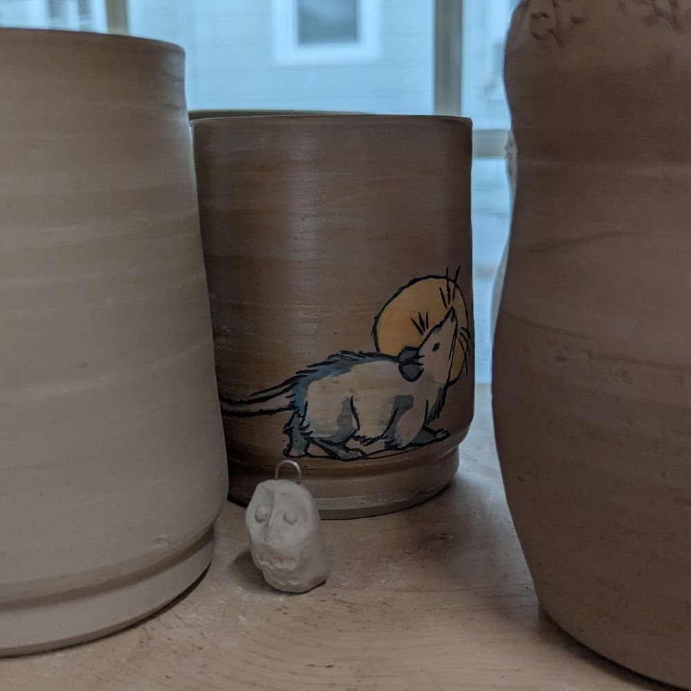 Unfired ceramic cups in a shelf with a small ceramic owl in front. One cup has a opossum in front of a moon or halo painted in underglaze.