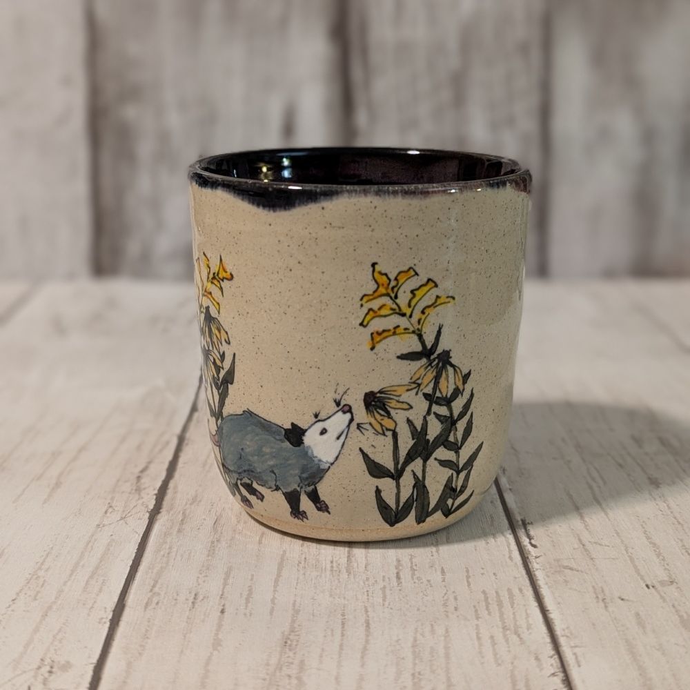 Ceramic cup with a painted opossum smelling black eyed susans and goldenrod flowers.