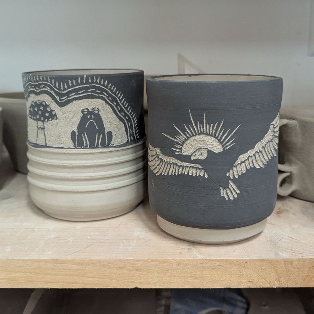 The cup on the left has a black and white carving on the top half of the cup with ridges in the clay on the bottom half. The carving is of a grumpy frog sitting next to a black and white Fly Agaric Mushroom. The cup on the right is a black ceramic cup with a vulture carved into it revealing the white clay under the black underglaze. The vulture has a sun halo behind its head. 