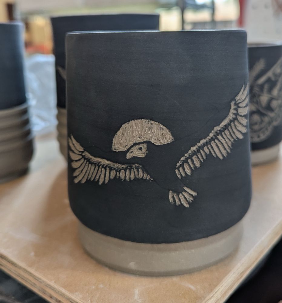 A black ceramic cup with a turkey vultures carved into the black underglaze to reveal the white clay underneath. 