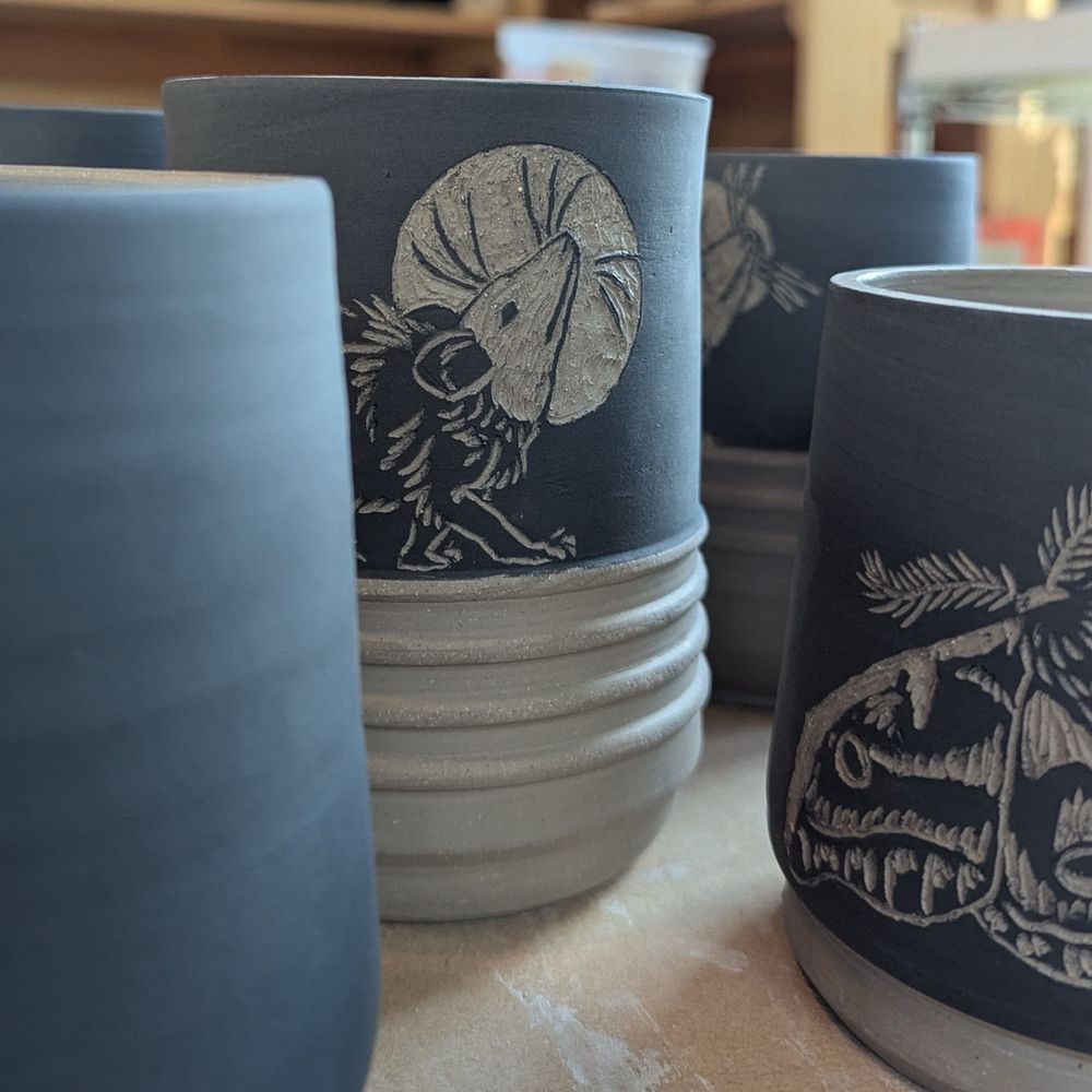 A board of 5 freshly carved ceramic cups. The top half of one has an opossum carved into black underglaze with its head up in front of a moon or halo. A second cup not in the center has a carved Polyphemus moth. 