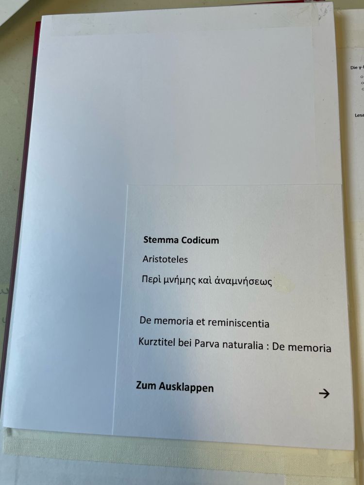 Aufklappbare Karte im vorderen Buchdeckel. Vorne ist aufgedruckter Text mit dem Hinweis, dass ein Stemma zu Aristoteles De Memoria et reminiscentia zum Aufklappen vorliegt. Auch der griechische Titel ist abgedruckt, unten ist ein Pfeil, um die Klapprichtung explizit zu zeigen.