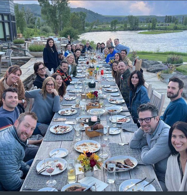 People outdoors at a long table near a body of water with various dinner dishes and beverage glasses in front of them.  Everyone at the table is White.