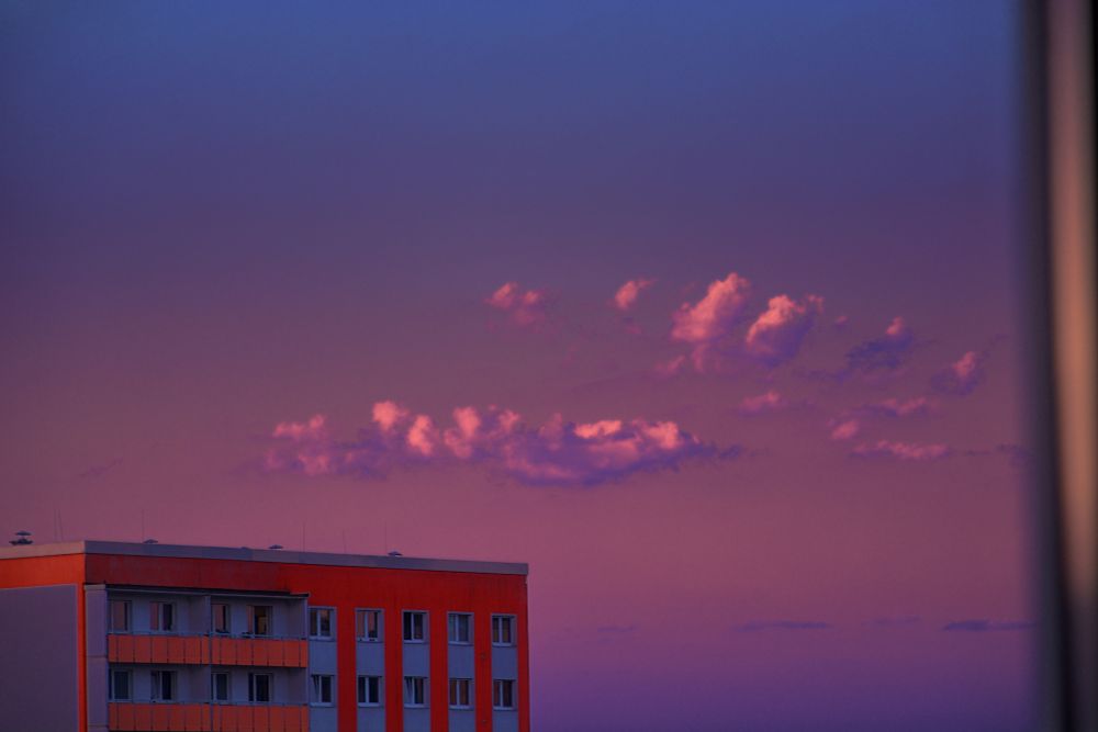 on the bottom left is the top part of a red-ish square, panel construction house. the main object of the photo are the pink clouds in the centre. top part of the photo is a gradient from pink to blue
