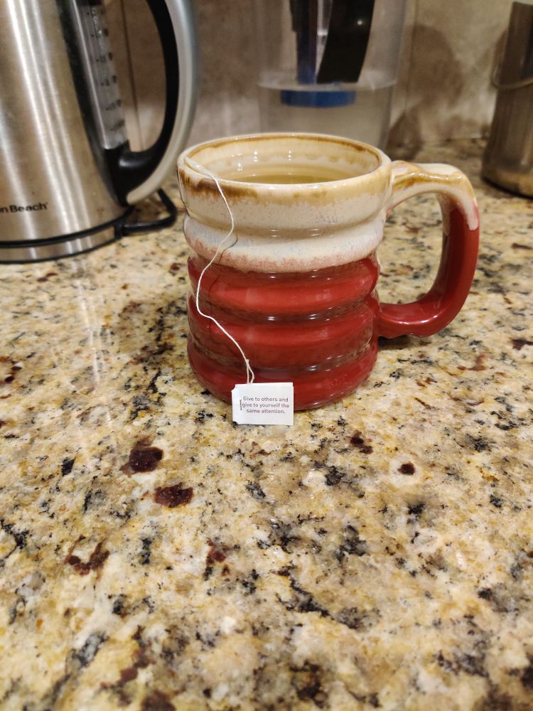 Red and tan ceramic pottery mug with Yogi Honey Chai Turmeric Vitality steep on a light granite countertop. The tag on the tea bag reads: Give to others and give to yourself the same attention.