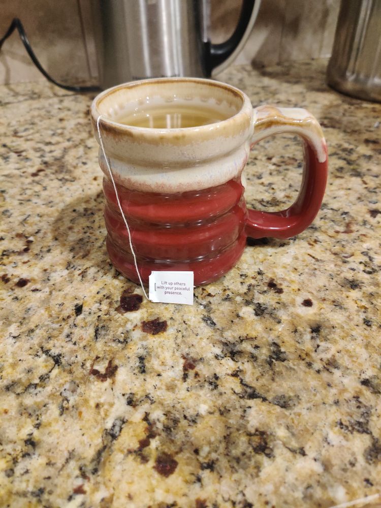 Red and tan ceramic pottery mug with Yogi Honey Chai Turmeric Vitality steep on a light granite countertop. The tag on the tea bag reads: Lift up others with your peaceful presence.