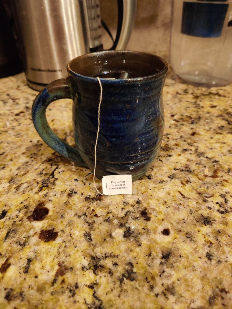 A dark blue pottery mug with some grey and brown on a granite countertop with Yogi Honey Chai Turmeric Vitality tea steeping. The tag reads: Forgiveness is an act of consciousness.