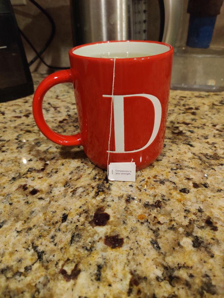 Tea (Yogi honey chai turmeric vitality) steeping in a large red mug with the letter D in a serif font on a light granite countertop. 

The tag on the teabag reads: Compassion is your strength.