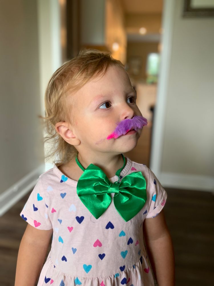 adorable caucasian toddler girl wearing green bowtie and colorful stickon mustache paired with pastel pink dress with heart pattern