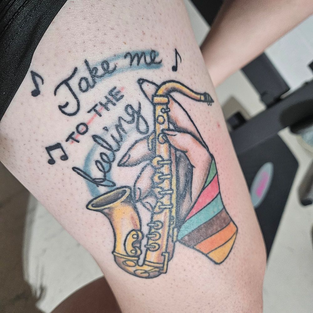 Tattoo on my thigh of a hand with a multicolored sleeve holding a saxophone with the lyric 'Take me to the Feeling" from Carly Rae Jepsen's song Run Away With Me. 