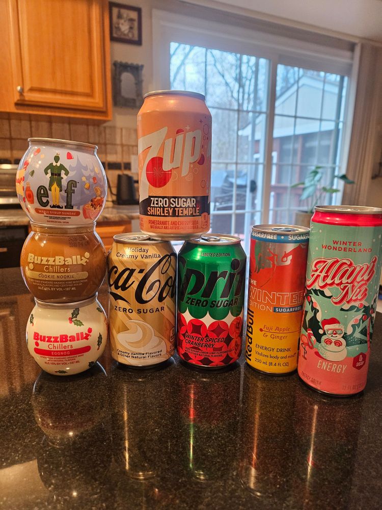 A bunch of various drinks in cans on a counter top.
Drinks include (from left to right)
-Buzzball Elf (Maple Syrup)
-Buzzball Cookie Nookie
-Buzzball Eggnog
-7Up Zero Sugar Shirley Temple
-Coca-Cola Zero Sugar Holiday Creamy Vanilla
-Sprite Zero Sugar Winter Spiced Cranberry
-Red Bull Sugar Free Winter Fuji Apple & Ginger
-Alani Nu Energy Drink Winter Wonderland (sugar plum flavored)