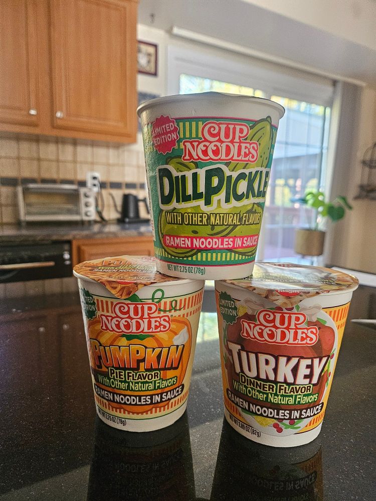 Three cups of CupNoodles Brand Instant Ramen. They are stacked in a pyramid formation. The flavors are Dill Pickle, Pumpkin Pie, Turkey Dinner.