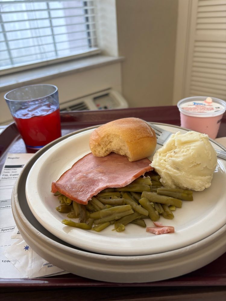 Plate with a scoop of mashed potatoes, overcooked canned green beans, slice of ham, roll missing a bite. Tray also has a cup of pink ice cream and a red drink with ice. 