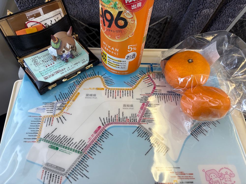 Satono Diamond -minifiguuri istuu matkalaukkukorttikotelossa allaan JR Shikokun reittikarttaa esittävä clear file -tasku ja katse kohti kartan Yosan Line -linjaa. Minifiguurin vieressä on mandariinin makuinen chuuhai-lonkero ja kaksi mandariinia