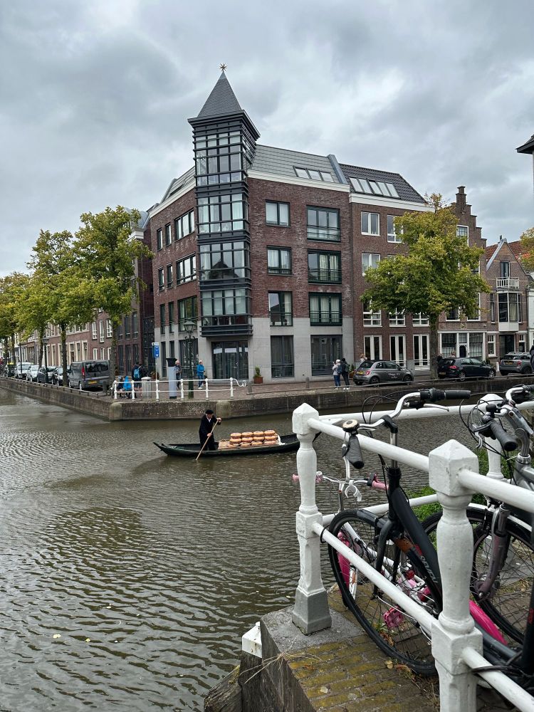 Um homem rema em um barquinho pelo canal em um dia cinzento, transportando uma pilha de queijos. Bicicletas estão estacionadas ao longo da calçada. Ao fundo, um prédio antigo de vários andares.