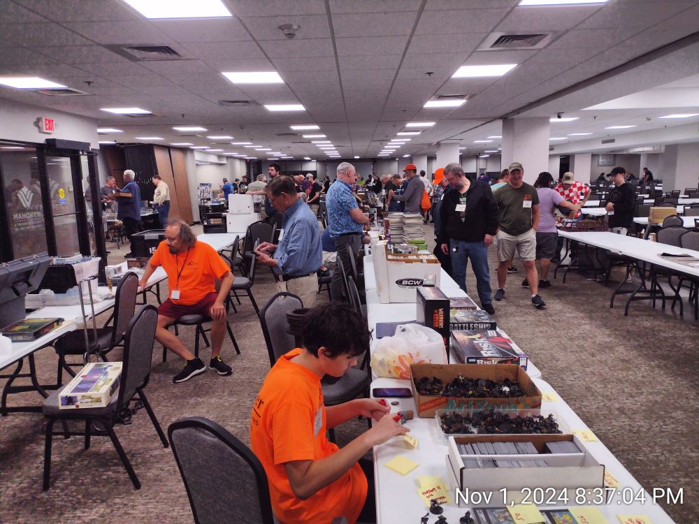 People standing and sitting around tables full of merchandise.  A few are wearing bright orange shirts, earned by volunteering at previous HMGS conventions