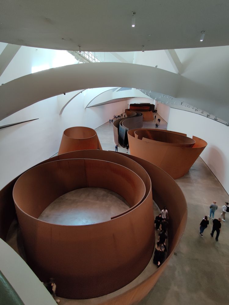 Riesengroße rostige Skulptur im Guggenheim-Museum in Bilbao. Die Skulptur stellt ein aus runden, ineinander geschachtelten Elementen bestehendes Labyrinth dar, betrachtet von oben. Darin bewegen sich Menschen, der umgebende Raum ist weiß mit gewölbten Bögen in der Deckenkonstruktion.