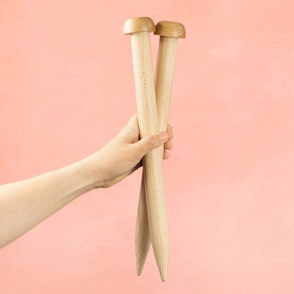 Large wooden knitting needles in someone's hand. Their diameter is almost the same as the length of the thumb. 