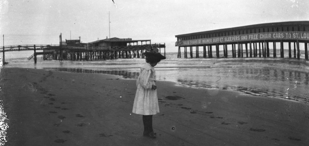 Tonight's historic Galveston photo is of an unidentified girl standing on the beach near the Pagoda Bathhouse (right), c. 1899. The structure in the background left is the Sea Breeze Saloon & Restaurant. Both those structures were wiped out in the 1900 Storm.

The reference to "Wagner Buffet Sleepers" on the Pagoda is part of the advertising for the Missouri, Kansas and Texas Railroad, colloquially known as "the Katy," that covered the structure. The Wagner Palace Car Company pioneered drop-down sleeper berths on rail cars, that were subsequently licensed to (and made famous by) the Pullman Car Company. Those berths weren't entirely comfortable, as I understand, but they were a vast improvement for long-distance rail travel over what had been the norm a generation before.

The girl is standing directly in front of the site of the famous Beach Hotel, that had burned in 1898, between 23rd and 24th Streets.
Rosenberg Library photo, SC285_100_2.