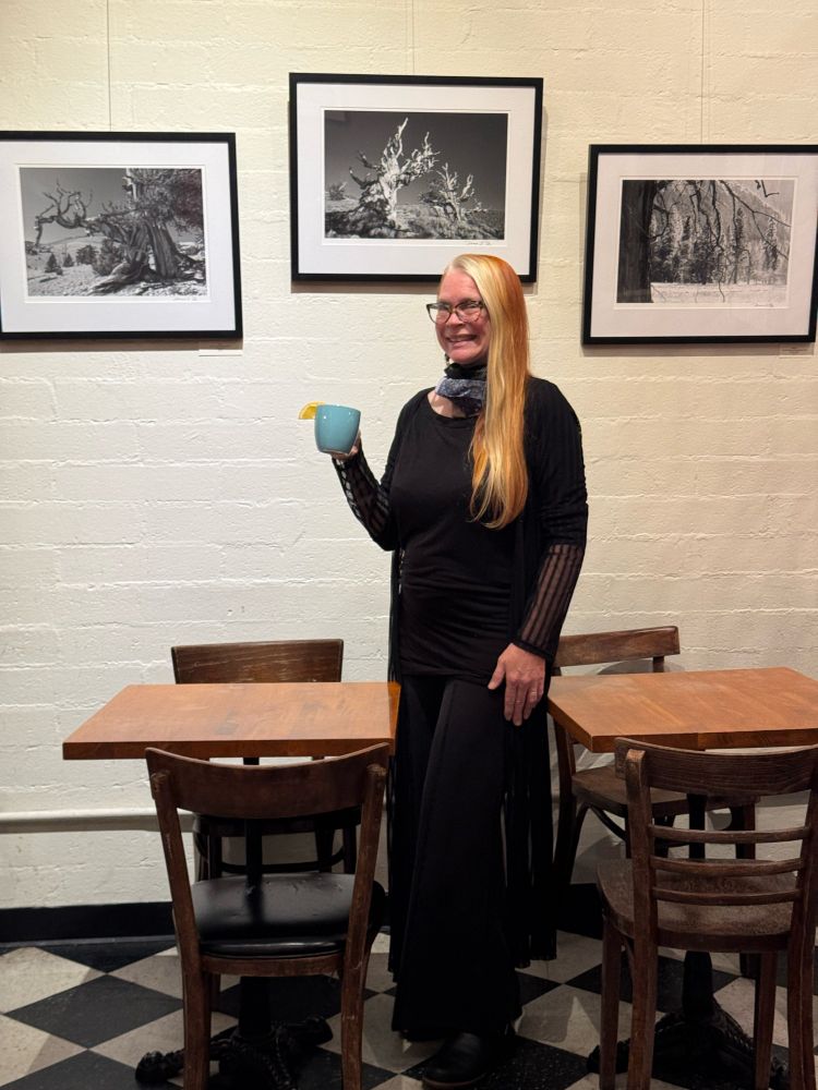 [Image ID: A photo of Denise standing in front of three of her black and white tree photos which are framed and on the wall behind her. There are brown wooden cafe tables and chairs on either side of her. She is holding a blue mug in her hand and smiling. End description.]