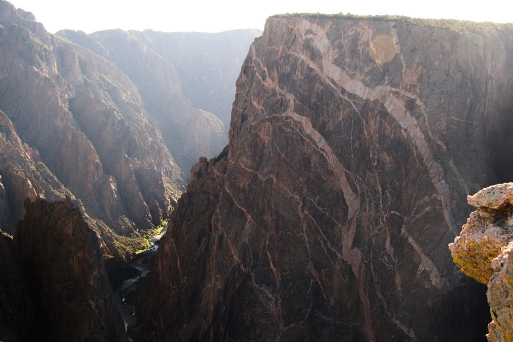 A photo of the 2250 foor shear cliff, the Painted Wall. It has veins of lighter rock across its face, and to the left of it is the river that carved the canyon, lit by some sunlight.