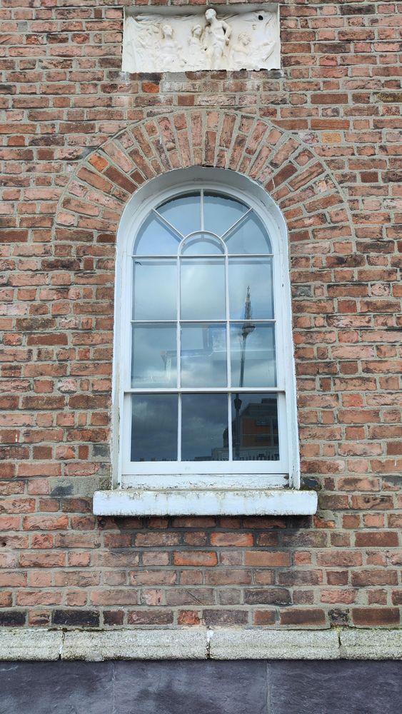 An arched window set in an old red brick wall. The cill is green with plant growth. Above the window a small rectangular white frieze is set into the wall