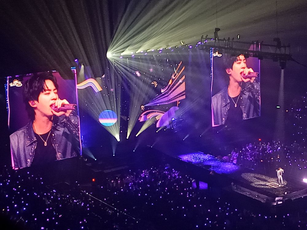 Kim Seokjin on the big screens during his first concert in London. He looks godly (as usual) wearing a jacket, black top, and necklace. He is holding his pink sparkly microphone to his mouth while singing, with his eyes shut. There are planet and stars motifs on the central stage screen, and the amibams in the crowd are flashing purple.