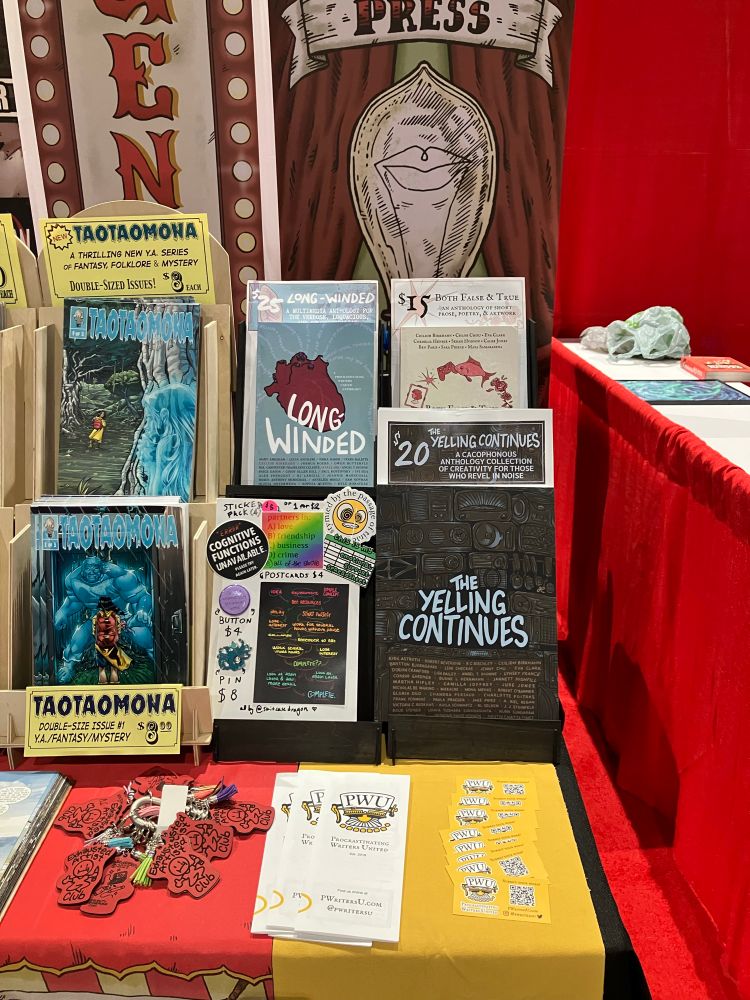 PWU’s booth setup, with all our inventory: physical copies of LONG-WINDED listed at $25, BOTH FALSE & TRUE at $15, THE YELLING CONTINUES at $20; a board with stickers, pins, and postcards, plus keychains, all by Ceilidh @suitcasedragon! As well as business cards and pamphlets.