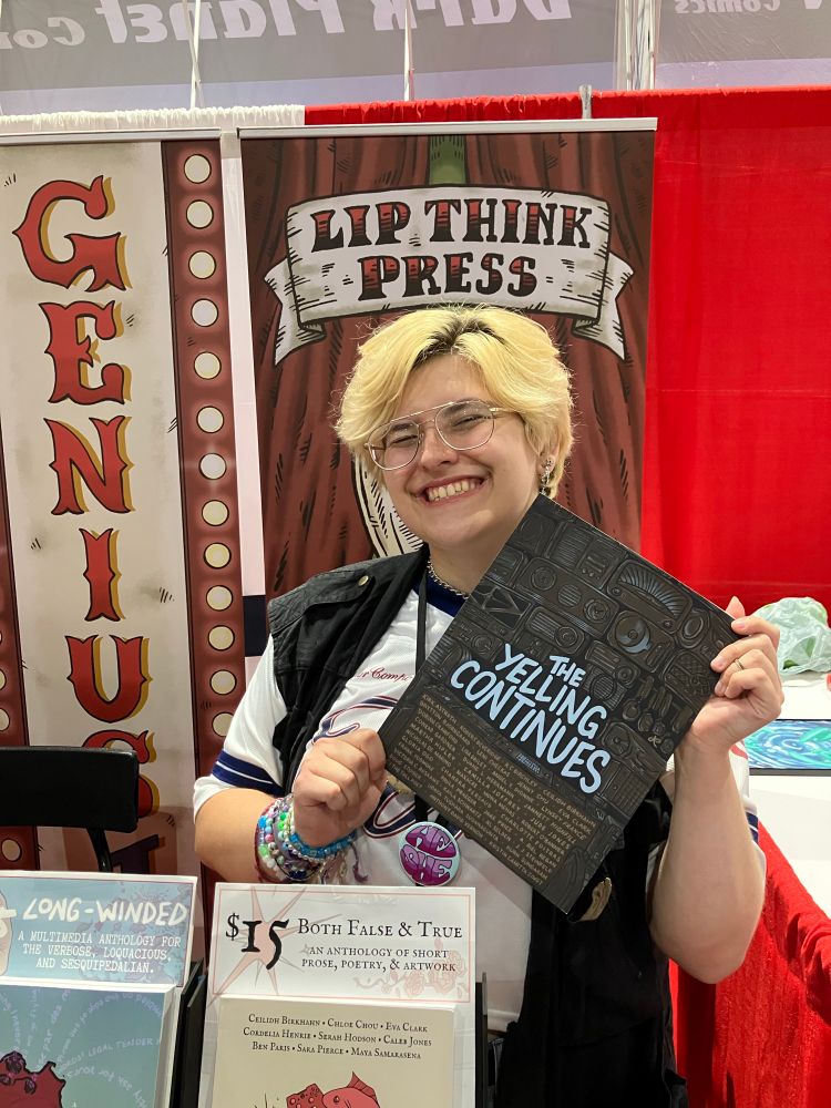 Eva holding a copy of THE YELLING CONTINUES in front of our publisher Lip Think Press’s banner!