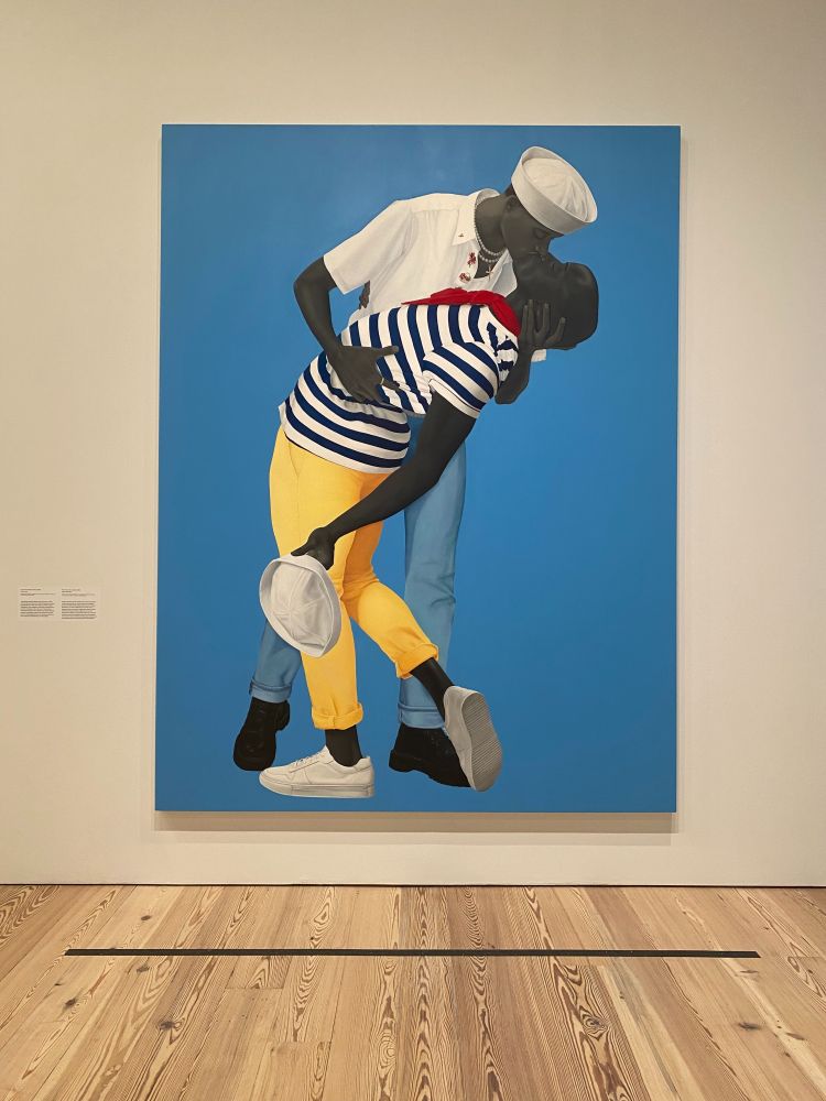 This is an image of a painting on a wall, showing an embrace of two men, kissing.  One is wearing a sailor hat and a pearl necklace, with a short sleeve shirt and jeans; he is supporting a man beneath him at his waist and behind his head,  in a striped nautical shirt and bright yelow paints, his foot kicked up precociously. 