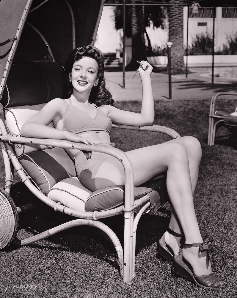 Lupino, in two-piece bathing suit, holding sunglasses, reclines in a lounge chair on a lawn with a pool and o palm trees visible in the background. 