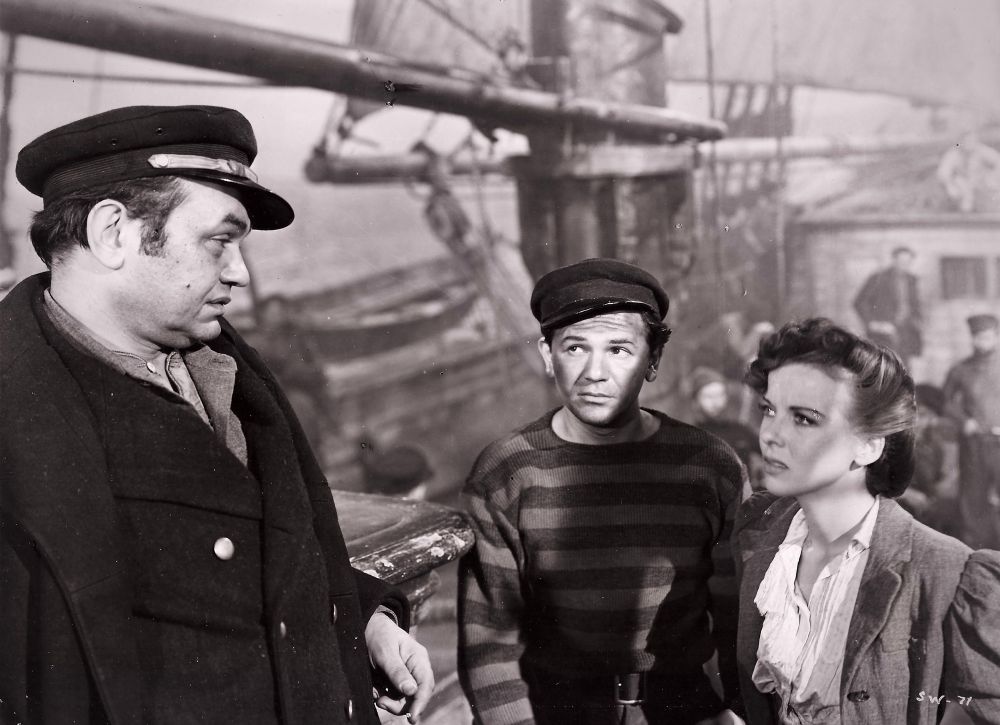 Robinson, Garfield and Lupino in costume, standing on deck of a large sailing ship. 