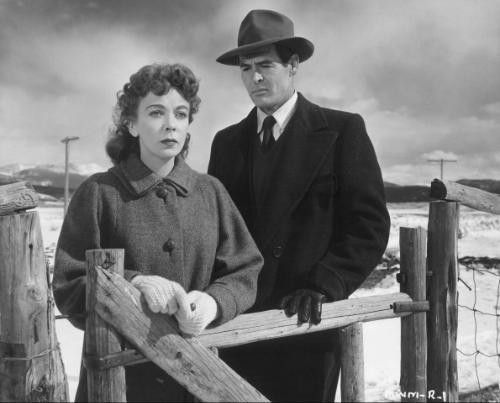 Lupino and Ryan stand solemnly by a wooden gate amidst a snowy landscape with mountains in the background. 