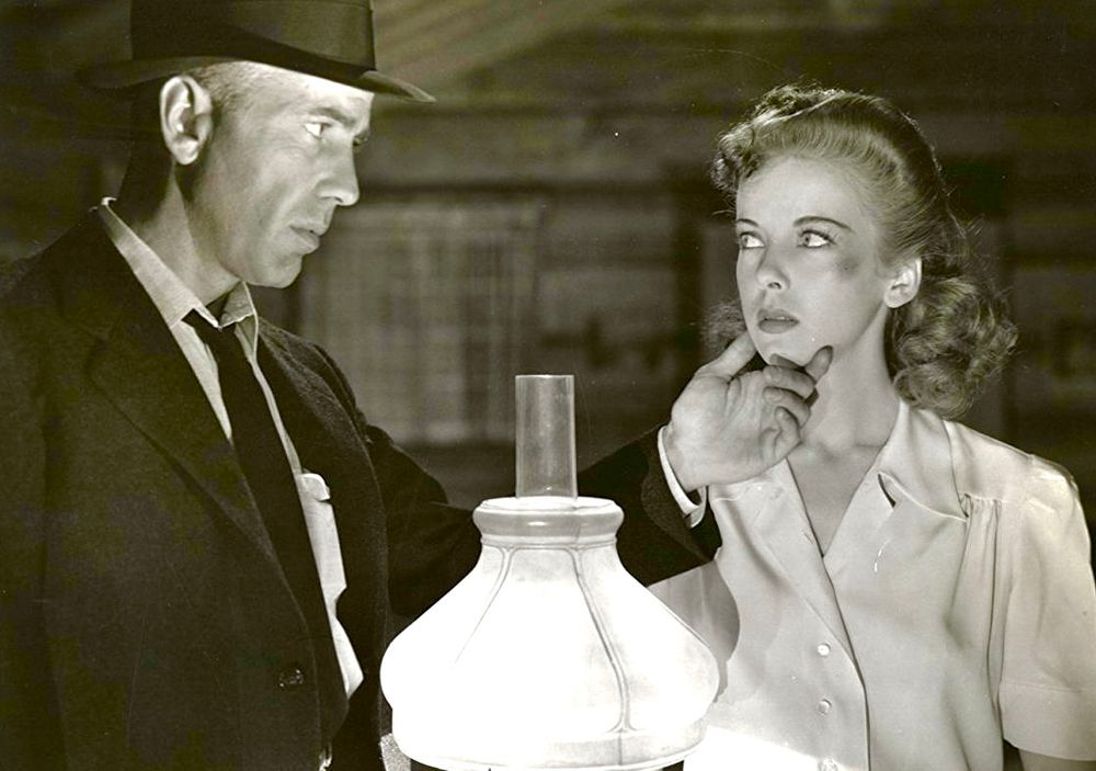 Beside a table with a lamp, Bogart holds Lupino’s chin to look at a bruise on her cheek near her left eye. 
