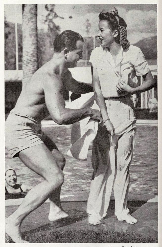 Lupino stands next to a swimming pool in soaked clothes. A muscular man in swim trunks leans in to her with a towel. 