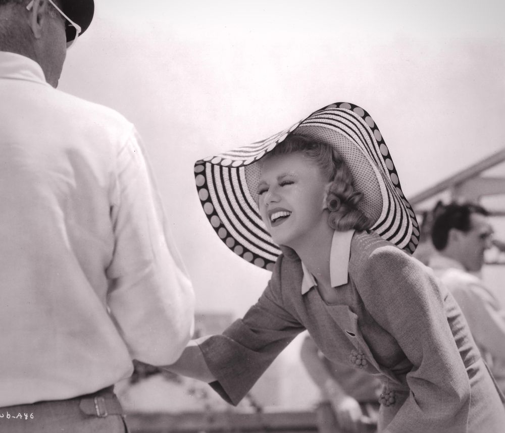 Ginger Rogers, outdoors in a fashionable hat with a very large brim, squints at LaCava, who has his back to camera at far left. 