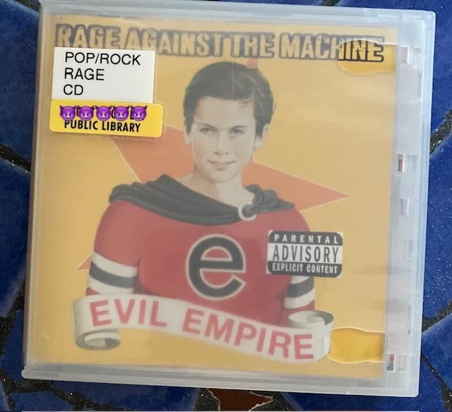 Album cover for Rage Against the Machine's Evil Empire. Yellow background featuring an illustration of Ari Meisel at 11 years old (according to Wikipedia), wearing a red, black and white costume with a lower-case "e" on the front. There are three stickers on the plastic case protecting the CD: 1) "Parental Advisory Explicit Content" 2) "POP/ROCK - RAGE - CD" and 3) "PUBLIC LIBRARY."  I've placed five purple demon emojis over the specific public library branch name. The background features blue rectangle and gold star mosaic tiles.  
