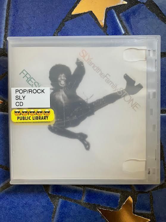 Image of the album cover for Fresh by Sly and the Family Stone, which features a gorgeous, joyful portrait of Sly Stone appearing to be in mid-air and wearing a fantastic outfit. There are two stickers on the plastic case protecting the CD: "POP/ROCK - SLY - CD" and "PUBLIC LIBRARY."   I've placed five sunglass emojis over the specific public library branch name. The background features blue rectangle and gold star mosaic tiles.