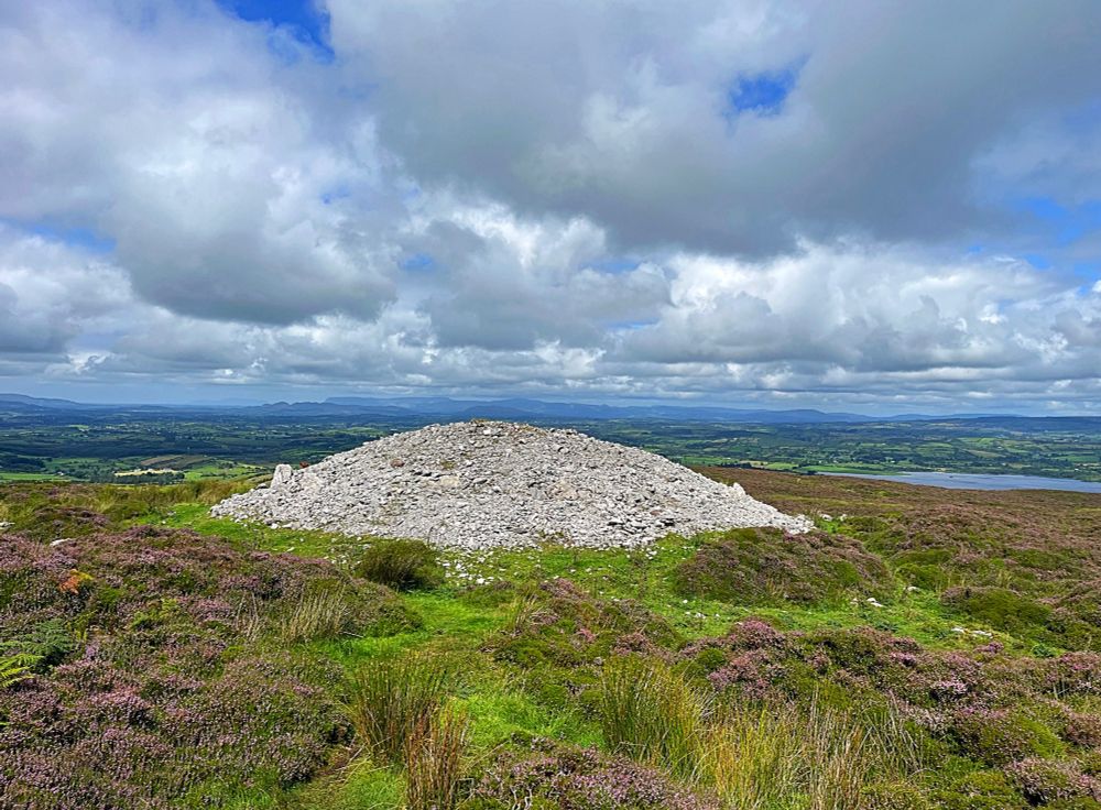 A photo of one of the Carrowkeel passage tombs. Grey stones forming a mound surrounded by green grass and purple heather.