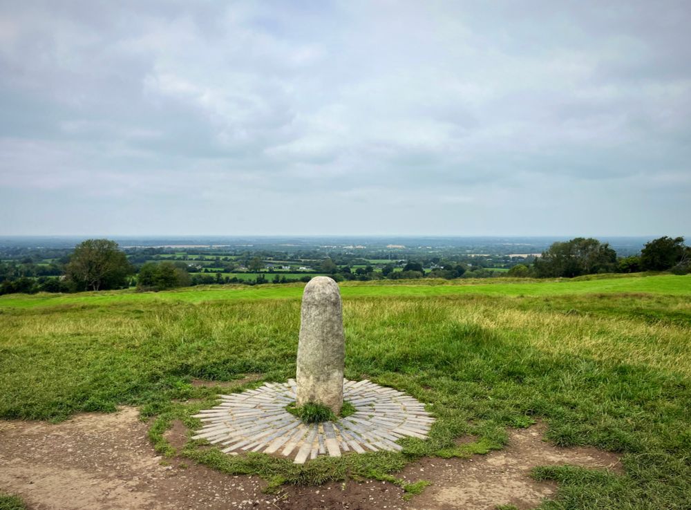 A photo of a pale pillar-like standing stone surrounded by a circular bricked area on grass in the foreground, with views out across the landscape in the background.