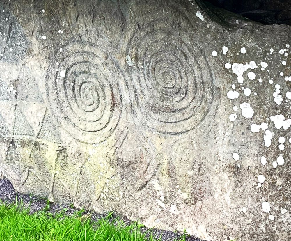 A photograph of a rock with the engraving of two spirals and a lozenge. The spirals form eyes and the lozenge a nose.