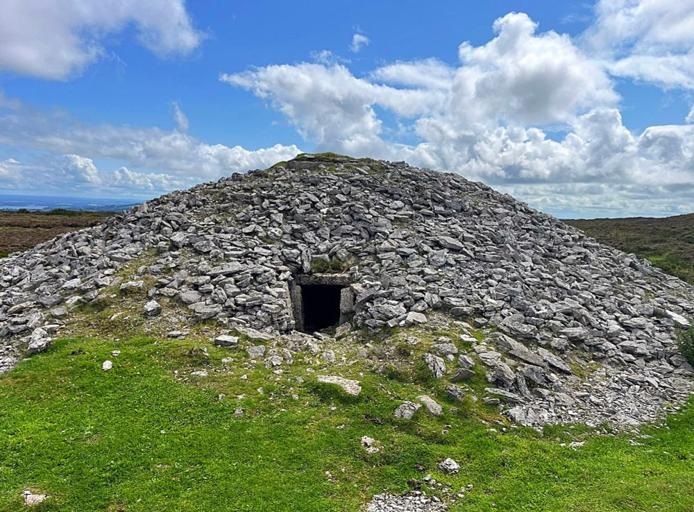 A photo of one of the Carrowkeel passage tombs. Grey stones piled over bright green grass and a doorway like a portal into the mound.
