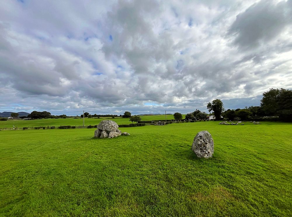 A photo showing a grey stone dolmen on green grass and with cloudy skies above. Another stone, a single surviving kerb stone is located slightly further away.