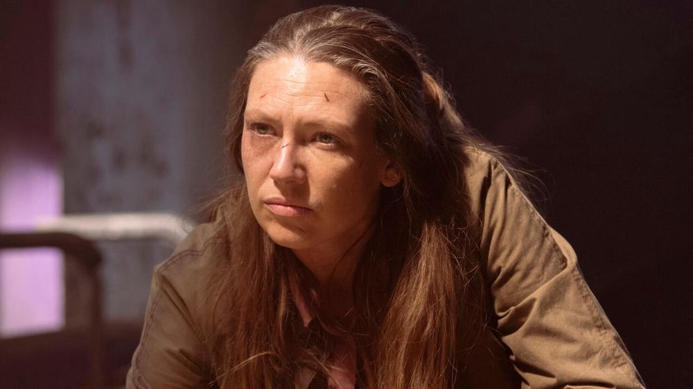 Anna Torv from The Last of Us season 2. Long Blonde hair with some grey in it that's pulled back from her face. She has small scrapes on her face and wearing a tan jacket.