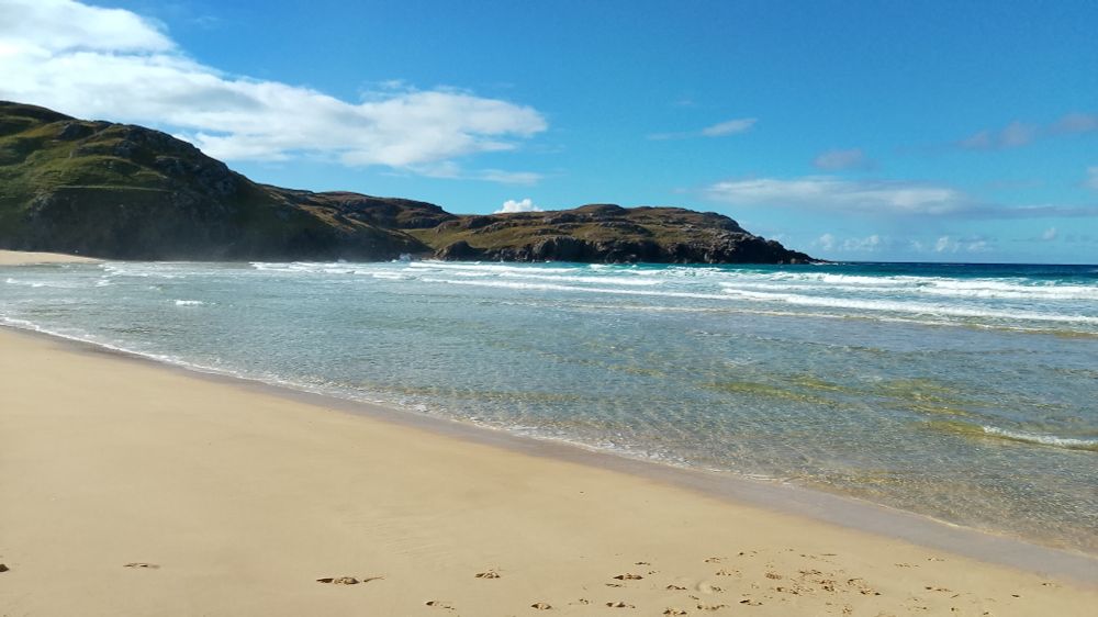 A glorious white/yellow sandy beach and clear azure sea against a low cliff backdrop in the Scottish Hebrides as the sun beats down.