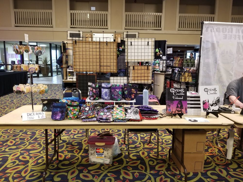Photo of a gaming booth at a convention. There are rpg books, dicebags and buttons for sale.