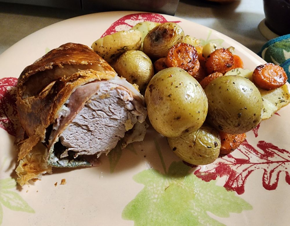A place with a couple slices of pork wellington and some roasted baby potatoes, carrots, and parsnips.