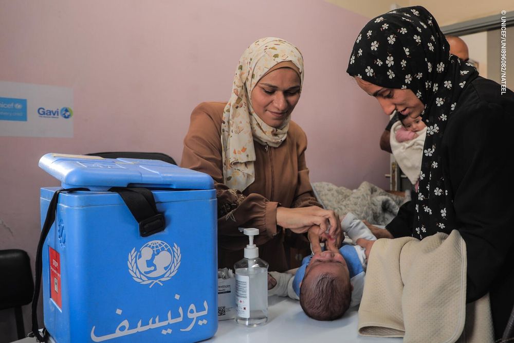 A health worker vaccines a child as a mother stands nearby. A cold storage vaccine container can be seen on the table.
