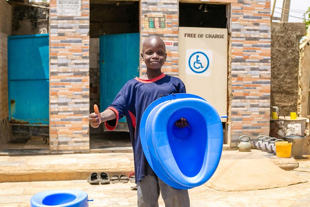 Saminu holds up a new toilet while giving a thumbs up. He's standing in front of a WASH facility.